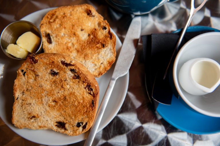 Teacake with teacup and milk A toasted teacake, open-side up on a plate with a butter knife, small metal pot of butter, empty teacup to the side with a small jug of milk