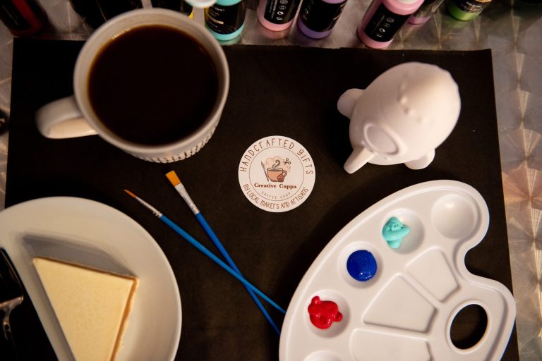 Painting activity whilst having coffee and cake. A white rocket ship, baby blue, dark blue and red paint set up with brushes for a customer with a coffee and a slice of cake to the side.